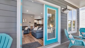 Une véranda couverte avec des fauteuils Adirondack de couleur turquoise, une porte-fenêtre coulissante donnant sur un salon avec un canapé gris et une cuisine en arrière-plan.