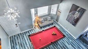 Een biljarttafel in een kamer met een sofa met zebraprint, een plafondlamp en een schilderij met een zebra-thema aan de muur.