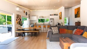 A modern living room with a grey sofa, wooden coffee table, and a dining area with a bench and table. - La Maison Soleil - Modern, sun-filled home with 4 bedrooms (2 kings including a stand-alone sleepout in the pool area, 2 queens) and 3 tiled bathrooms. Features a full kitchen, aircon, and open living flowing to a large garden with BBQ, heated swim s (Waiheke Island)