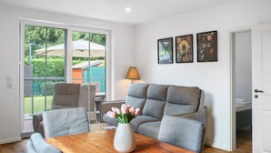 Una sala moderna con un sofá gris, una mesa de centro de madera y un jarrón con flores. Hay dos cuadros en la pared y una puerta que da a otra habitación.