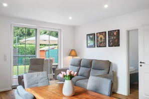 A modern living room with a grey sofa, a wooden coffee table, and a vase of flowers. There are two framed pictures on the wall, and a door leading to another room.