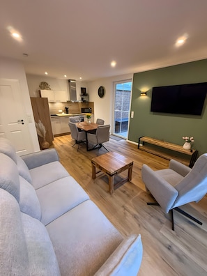 A modern living room with a sofa, coffee table, and armchairs, a dining area with chairs and a table, a kitchen with cabinets and appliances, and a TV mounted on the wall. - Holiday Home 'Thiesa' with Private Garden and Wi-Fi (Berumbur)