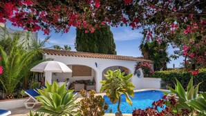 A pool area with lounge chairs, a white umbrella, and a building with arches. - Hibiscus by Sun & Sea Homes (Calpe)