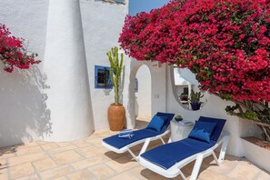 Un espace extérieur lumineux, aménagé pour se détendre, avec des fauteuils blancs et des coussins bleus, entouré de murs blancs et de magnifiques fleurs de bougainvillier roses.
