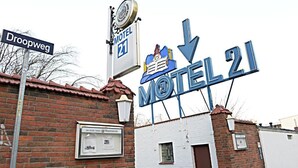 Un letrero de motel con una flecha hacia abajo, una señal de calle que dice 'Droopweg' y un pequeño edificio con fachada de ladrillo.