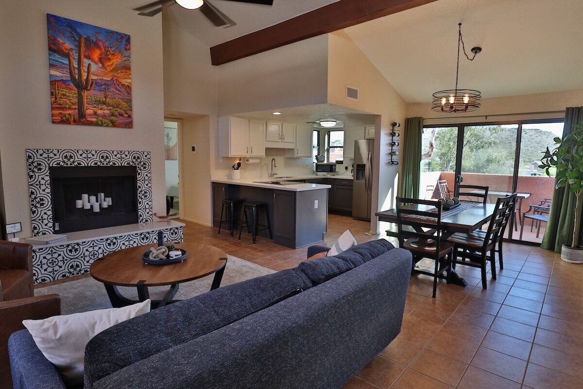 A living room with a fireplace, a sofa, a dining area with a table and chairs, and a kitchen in the background.