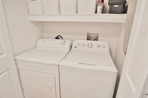 Une buanderie avec deux machines à laver blanches et une étagère au-dessus, où sont rangés des vêtements pliés et une bouteille.