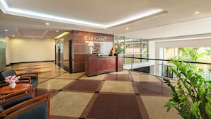 A lobby with a reception desk labeled "ELEGANT," a checkered floor, a seating area with a table and chairs, and a view through a glass wall.