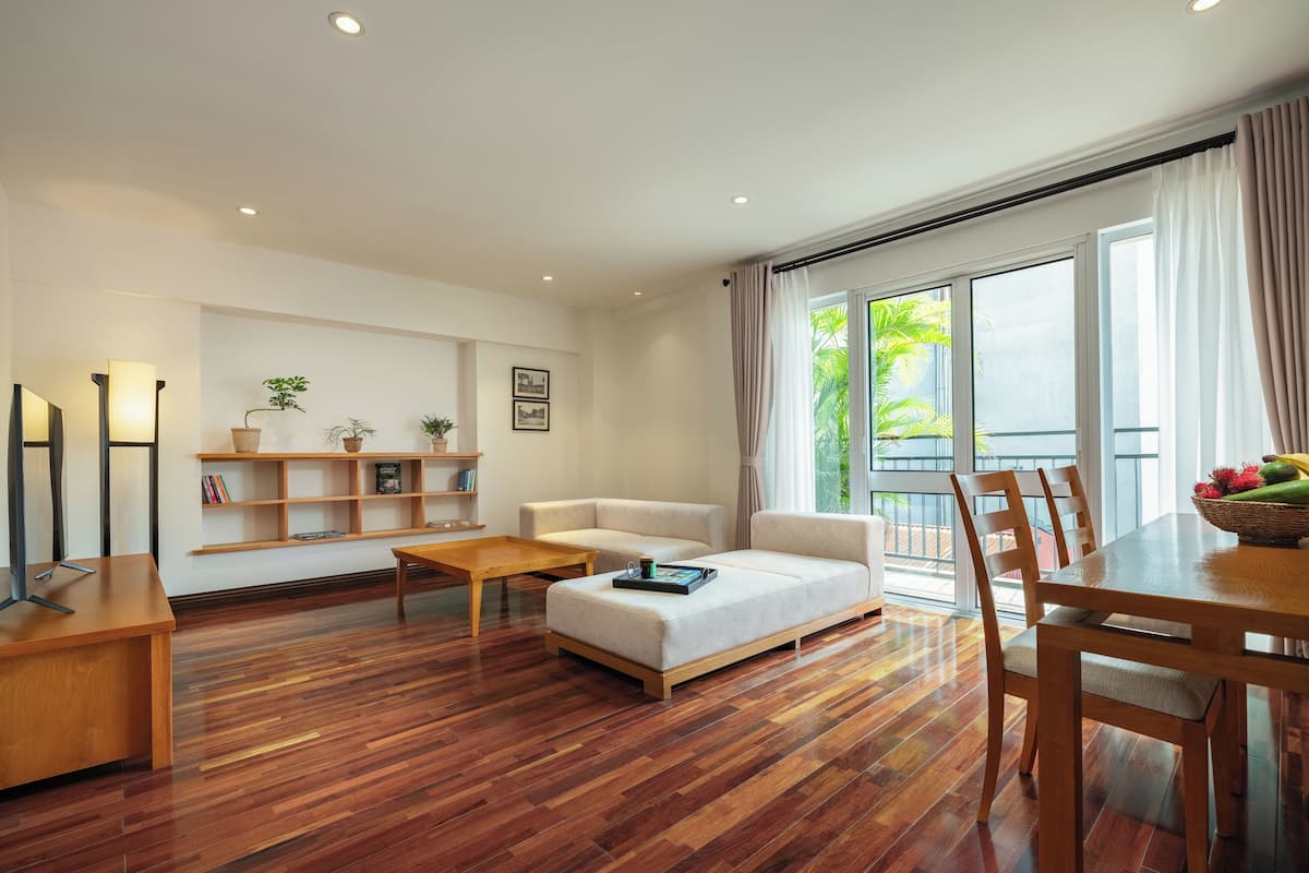 A modern living room with a wooden floor, a sofa, a coffee table, a dining table with chairs, and a bookshelf with plants.