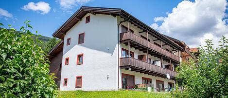 Un immeuble à plusieurs étages, doté de balcons et d’une façade blanche, avec une chaîne de montagnes en arrière-plan.