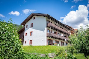 A multi-story building with balconies, a white facade, and a mountainous backdrop.