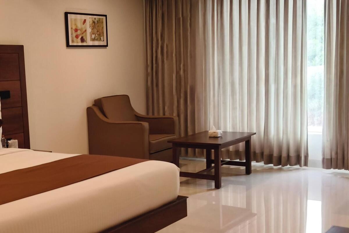 A hotel room with a large bed, a wooden headboard, a brown armchair, a small table, and a window with curtains.