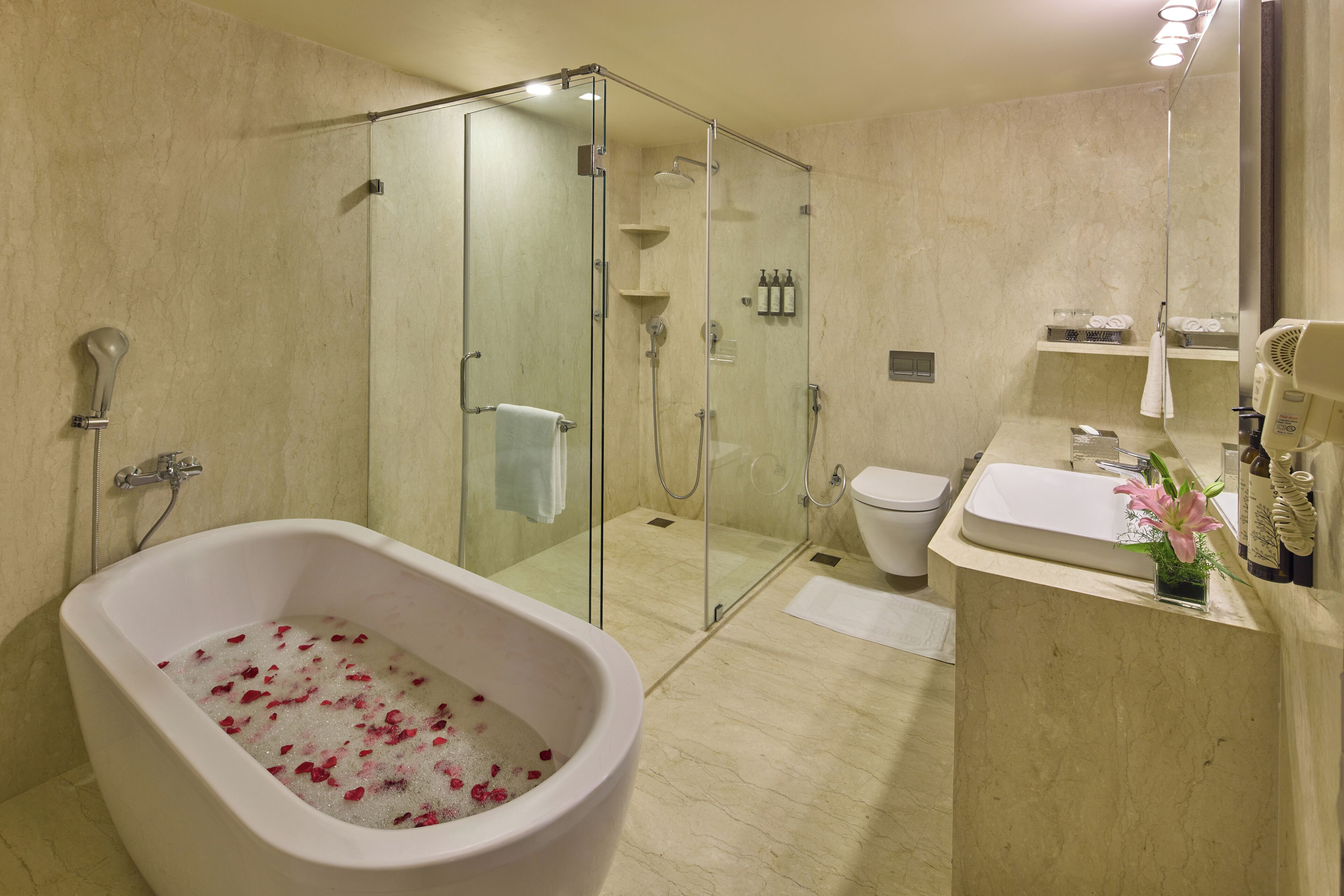 Une salle de bain moderne avec une baignoire sur pieds remplie d’eau et de pétales de roses, une douche à l’italienne, des toilettes et un lavabo agrémenté d’une petite plante.