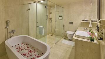 Un baño moderno con una bañera independiente llena de agua y pétalos de rosa, zona de ducha con mampara de vidrio, inodoro y lavabo con una pequeña planta.