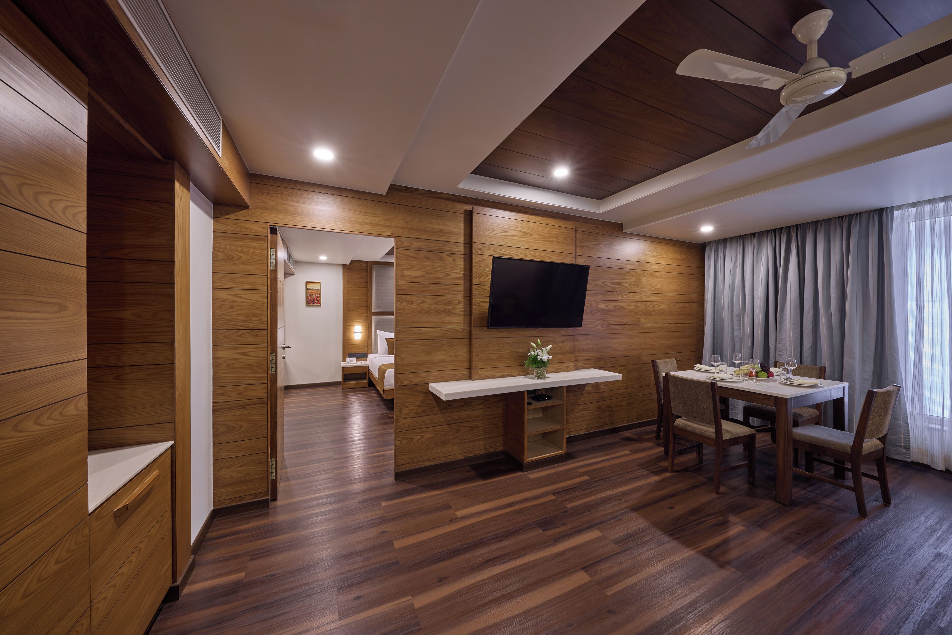 Una habitación de hotel moderna con piso de madera, un comedor con mesa y sillas, un televisor de pantalla plana en la pared y un ventilador de techo.