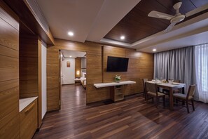 A modern hotel room with a wooden floor, a dining area with a table and chairs, a flat-screen TV mounted on the wall, and a ceiling fan. - Regenta Hubbali (Hubli)