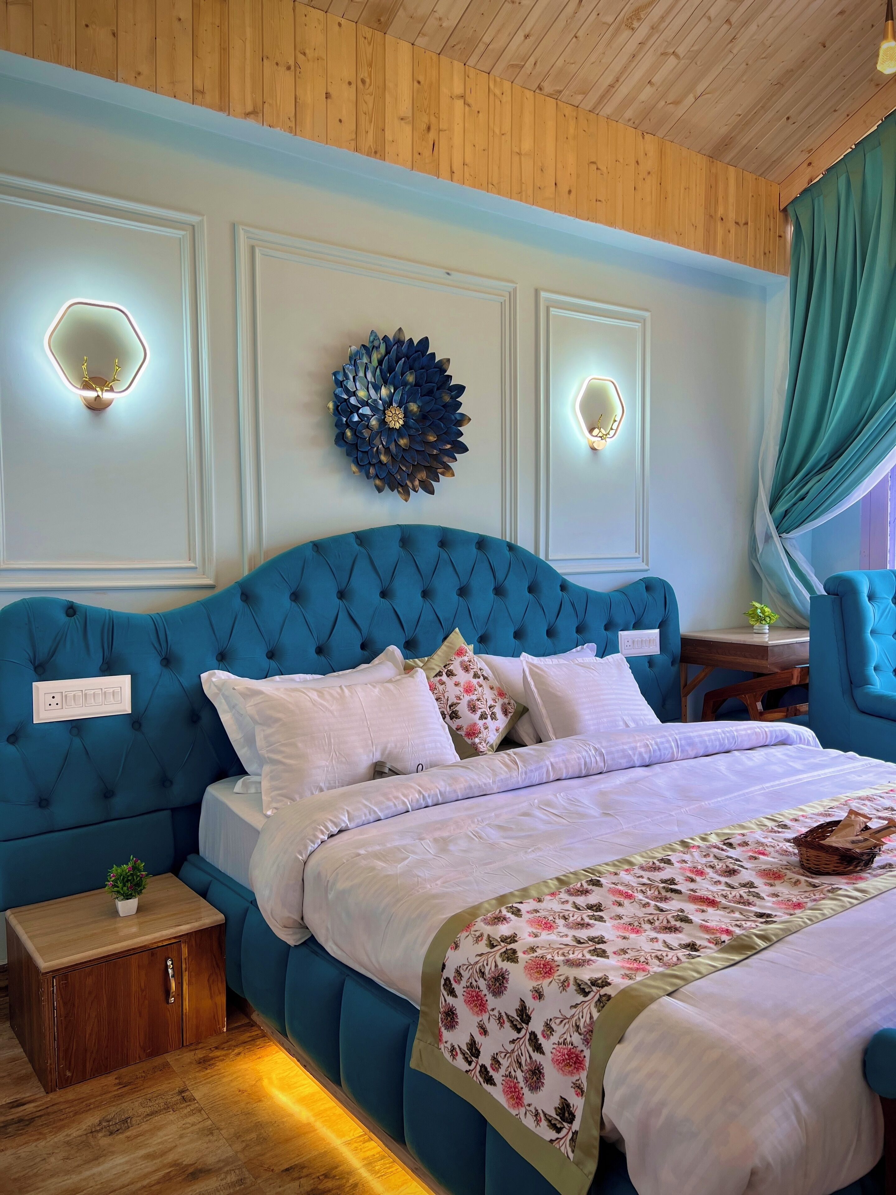 A bedroom with a blue tufted headboard, a floral bedspread, and two wall-mounted light fixtures.