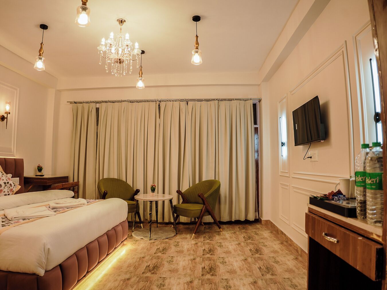 Quarto de hotel com cama, duas cadeiras, uma mesinha, televisão e um candelabro.