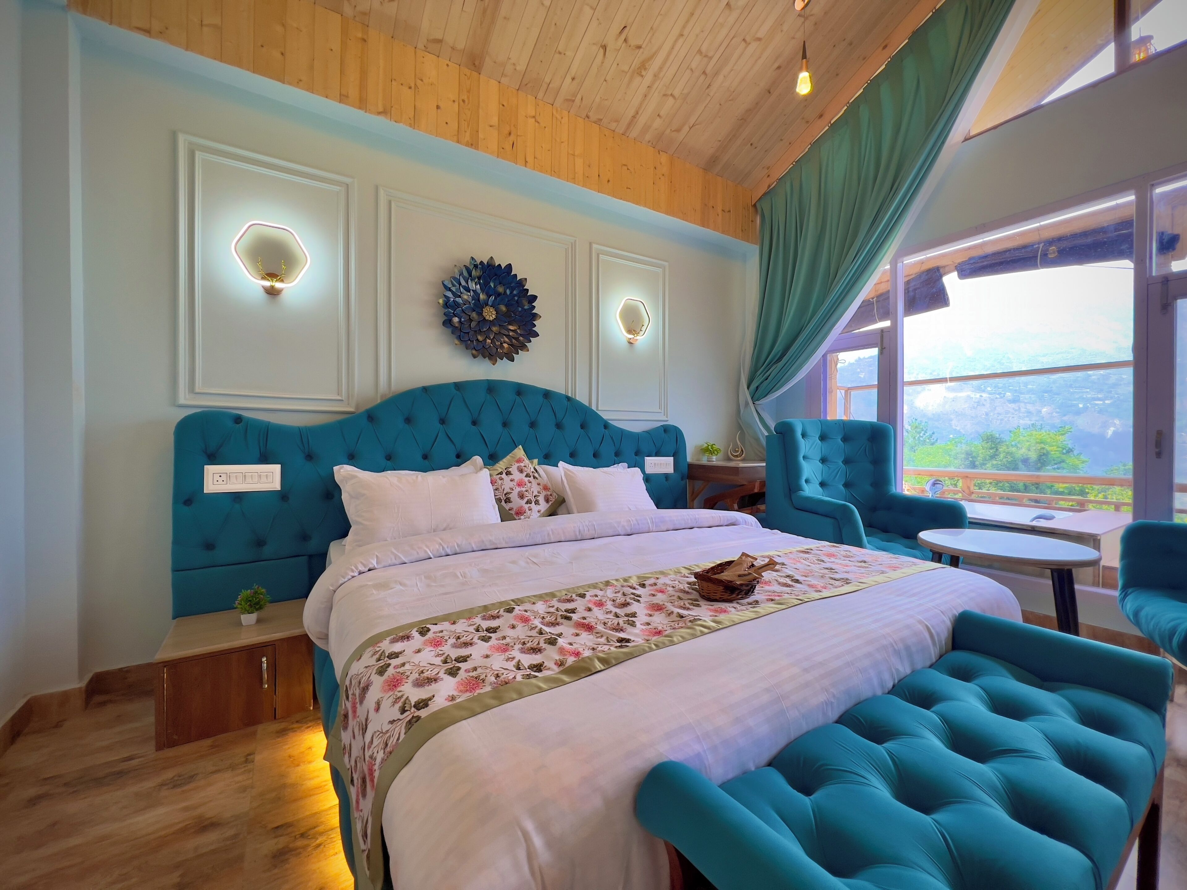 A bedroom with a large bed, a wooden ceiling, a view of the outdoors, and a small table with chairs.