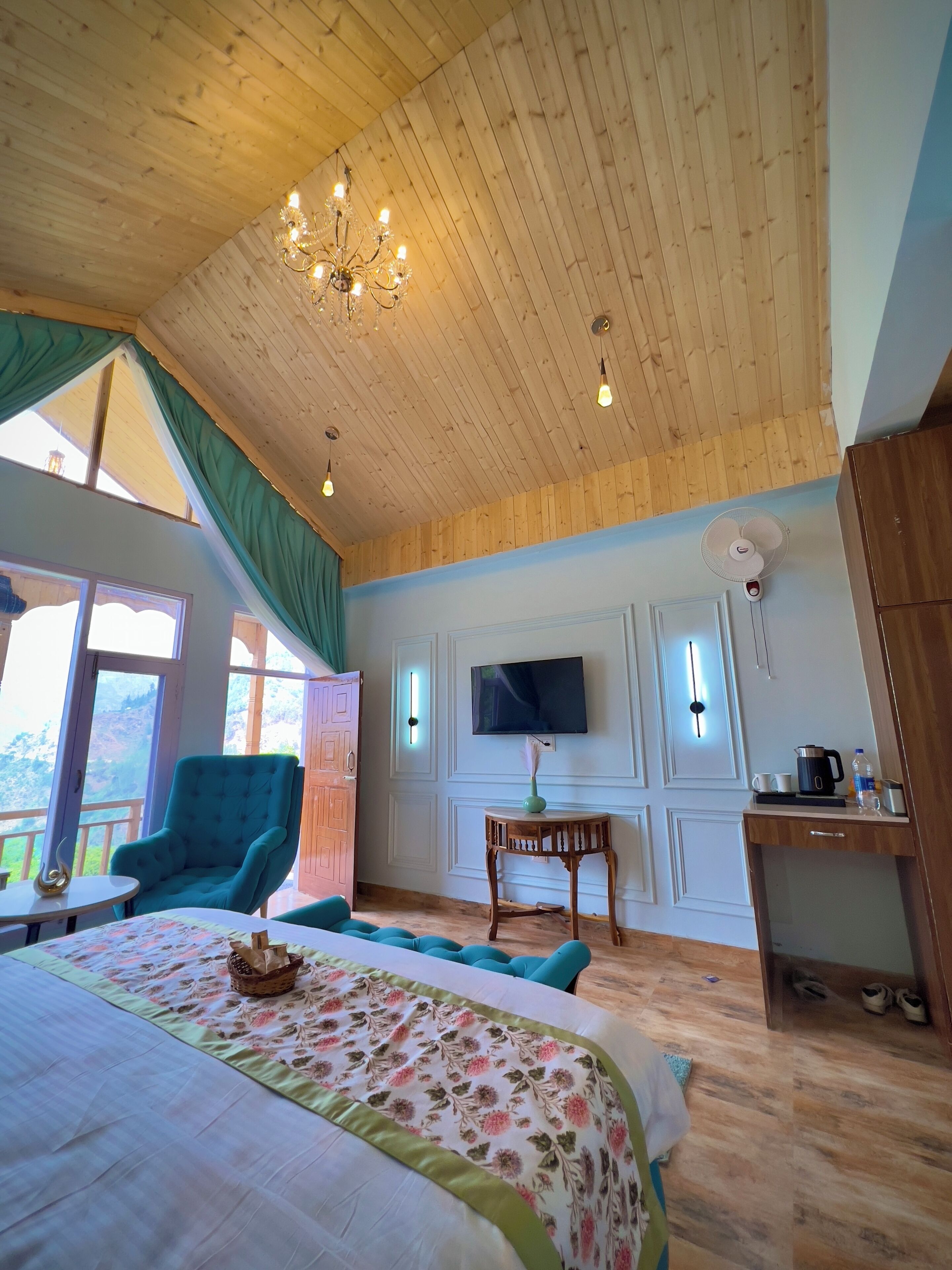 Quarto com teto de madeira, cama com roupa de cama florida, poltrona verde, pequena mesa com um vaso e televisão na parede.