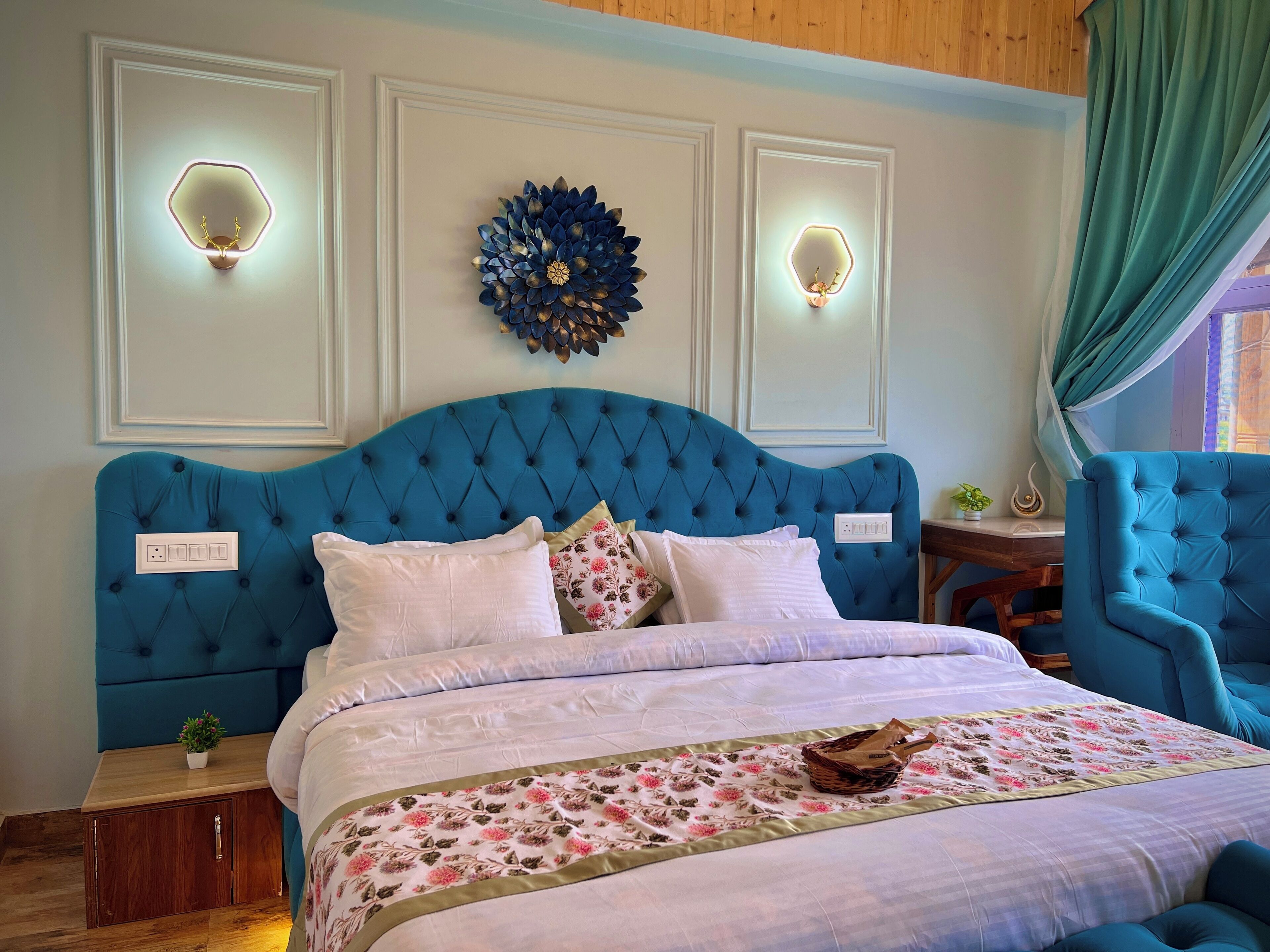A bedroom with a blue upholstered headboard, a floral-patterned bedspread, and decorative wall sconces.