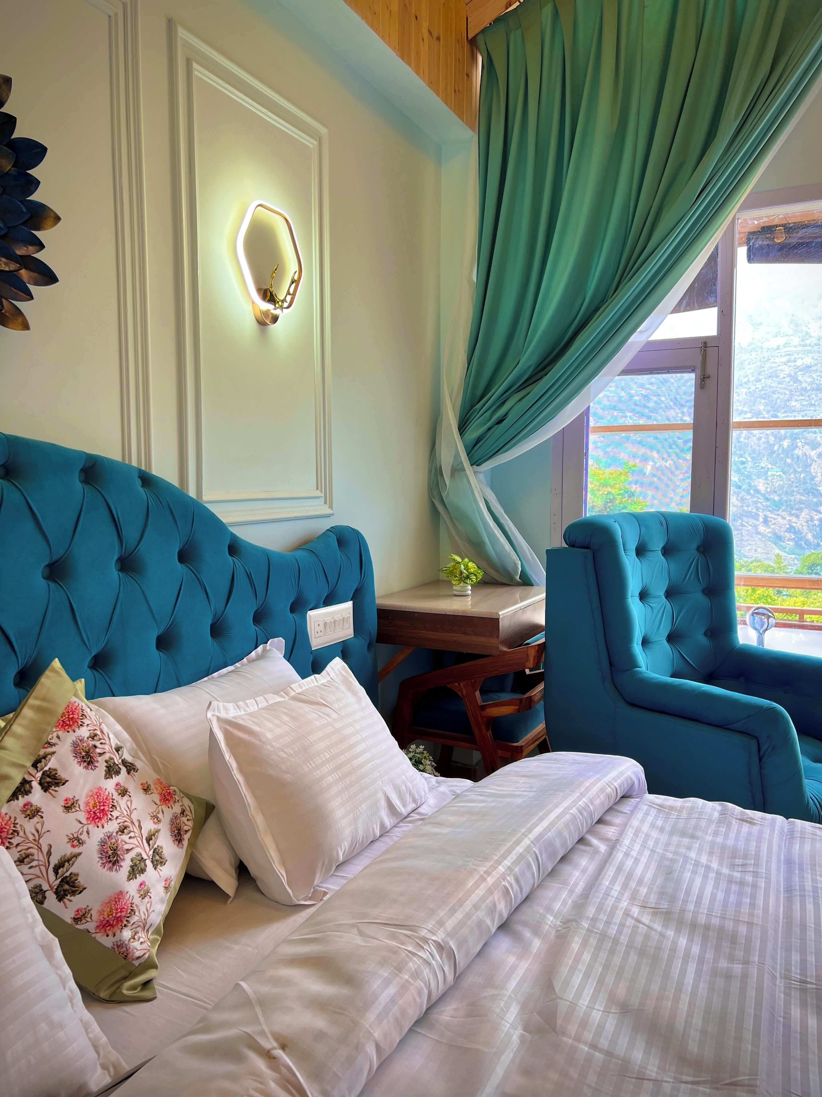 A bedroom with a tufted headboard, a bedside table with a lamp, a chair, and a view of greenery outside.