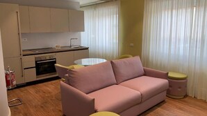 Een compacte leefruimte met een kitchenette, een roze bank, een klein rond tafeltje en een gele kruk.