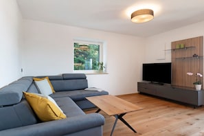 Una sala de estar moderna con un sofá seccional gris, una mesa de centro de madera, un televisor de pantalla plana y un ventanal con vista a zonas verdes.