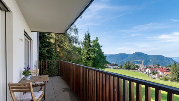 A balcony with a wooden table and chairs, a view of mountains and a village below.