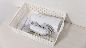 En hvid plastikkurv med en Panasonic hårtørrer, en hvid ledningsbaseret hårtørrer og en hvid elektronisk enhed med digital skærm.
