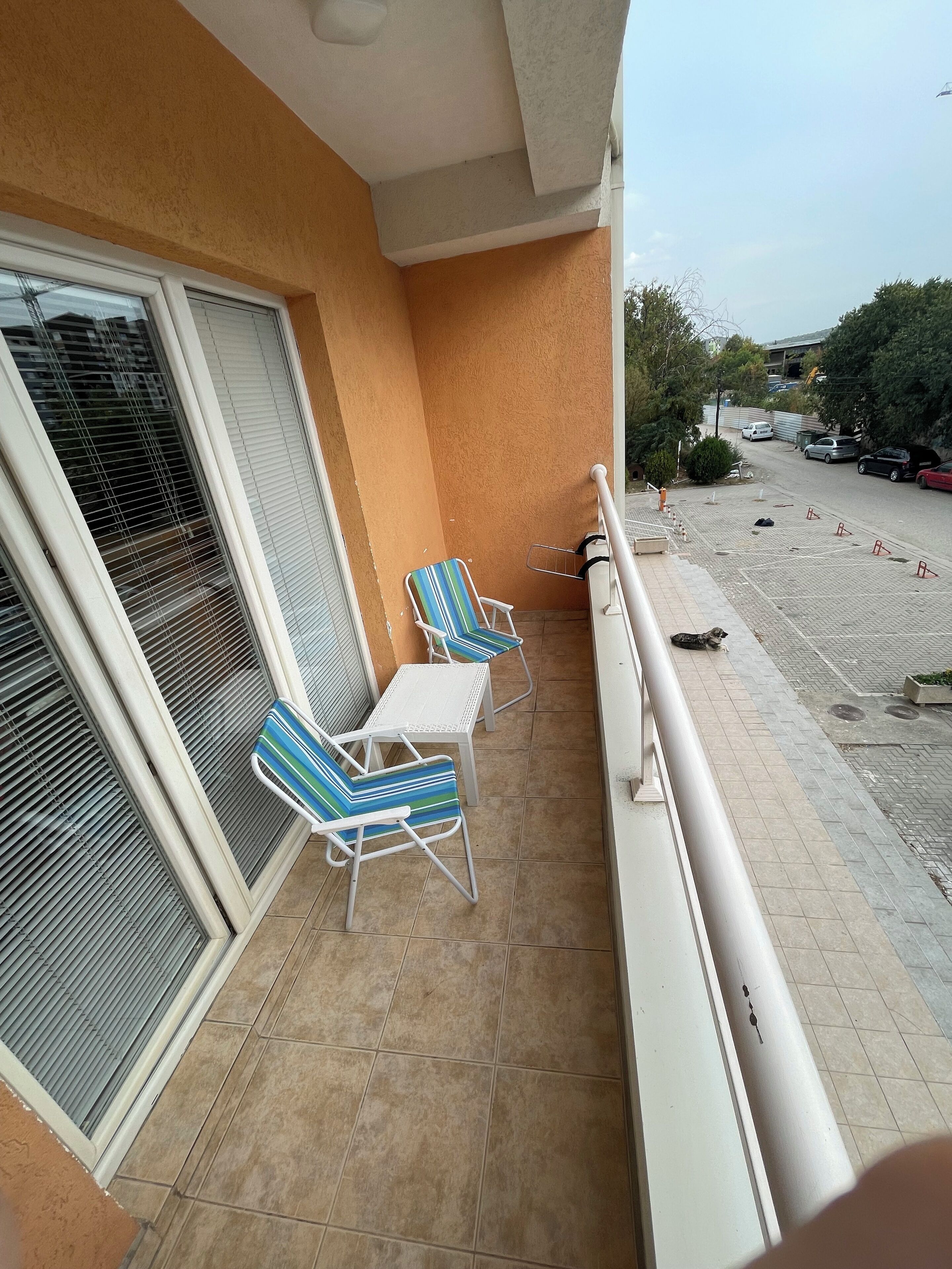 A balcony with two striped chairs and a small table, a railing, and a view of the street with parked cars and construction.