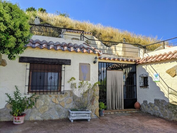 Un bâtiment avec un toit en tuiles, des murs en pierre, une fenêtre grillagée et des plantes en pot.