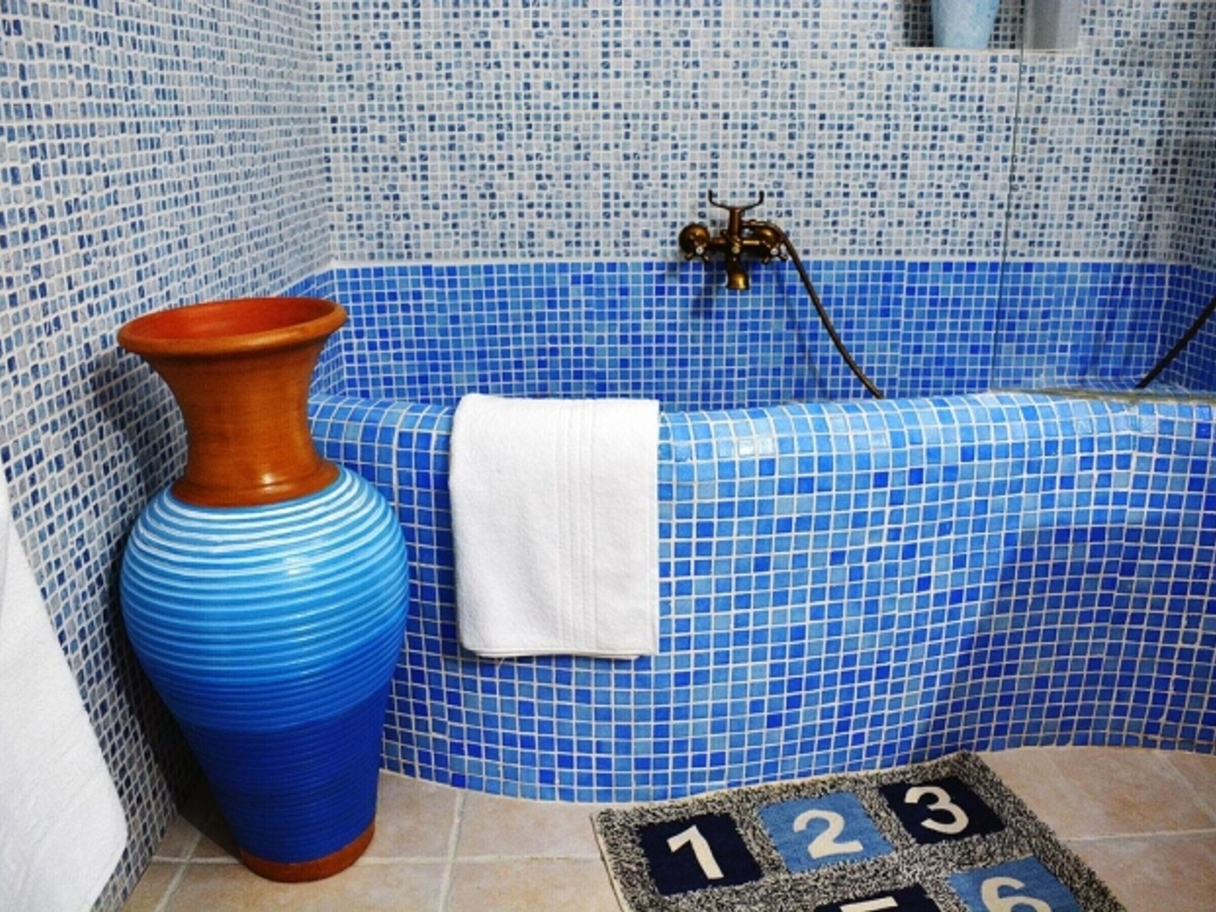 A bathroom with a blue tiled bathtub, a white towel, and a blue vase.