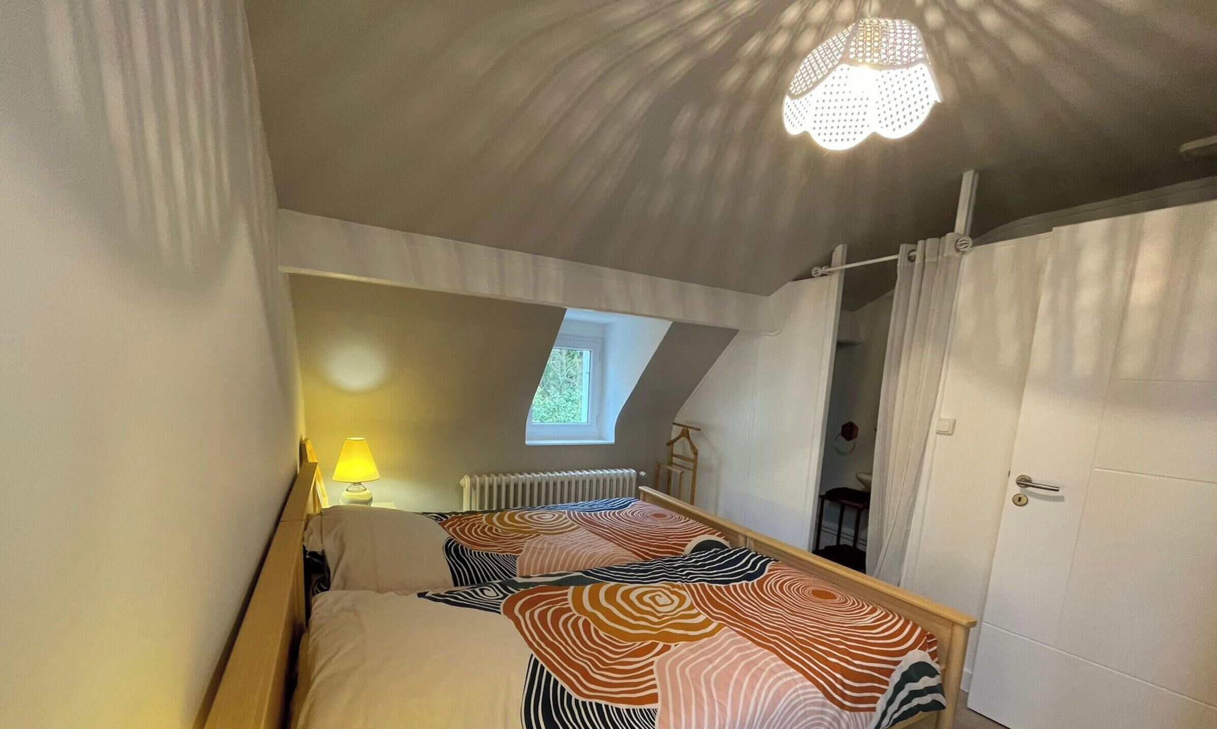 Ein Schlafzimmer mit geneigter Decke, einem Bett mit gemustertem Überwurf, einem Fenster, einem Heizkörper und einer Tür.