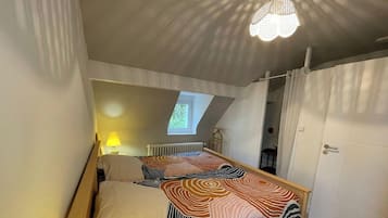 Ein Schlafzimmer mit geneigter Decke, einem Bett mit gemustertem Überwurf, einem Fenster, einem Heizkörper und einer Tür.