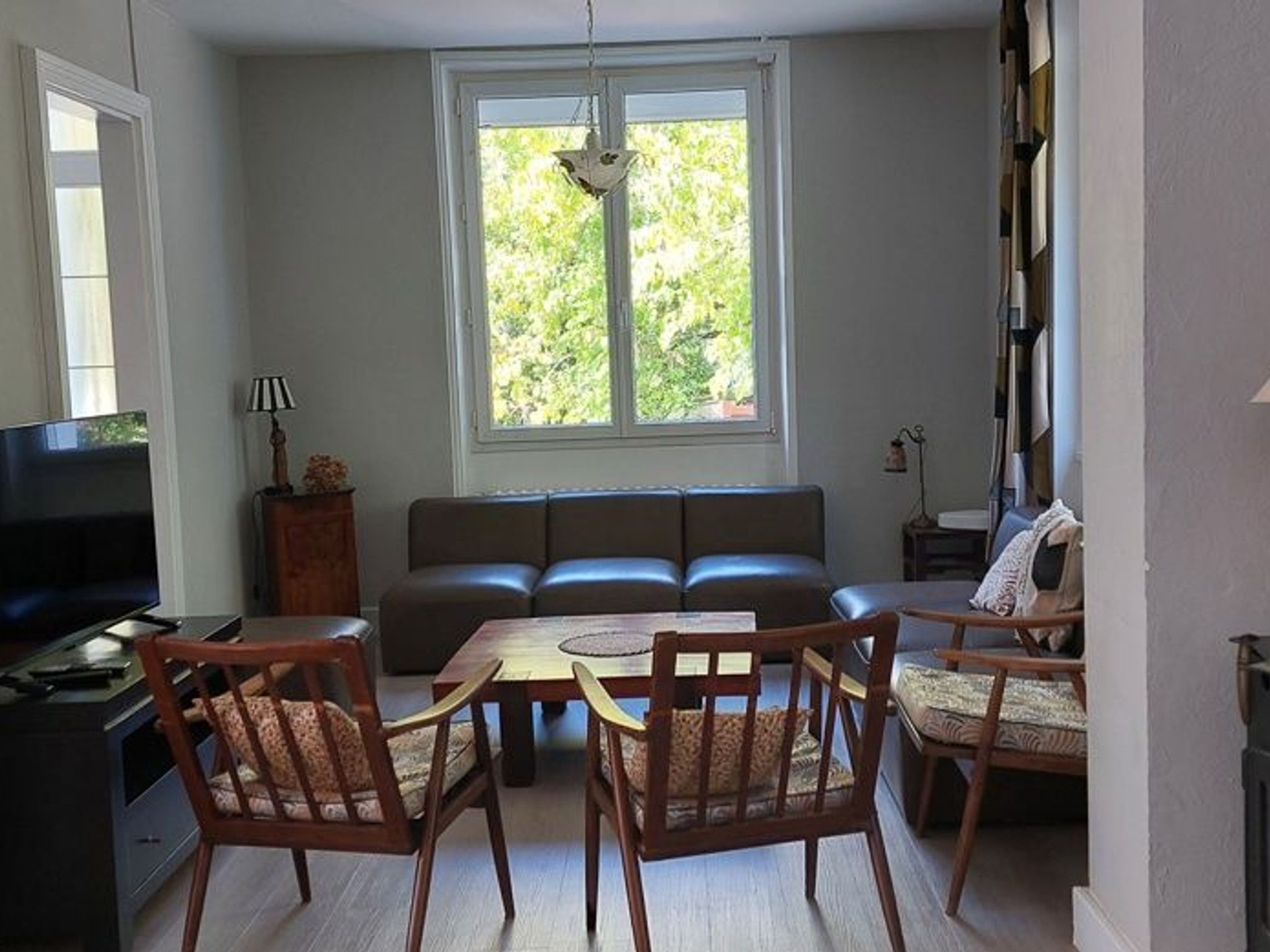 Een woonkamer met een bank, twee stoelen, een televisie en een raam met uitzicht op groen.