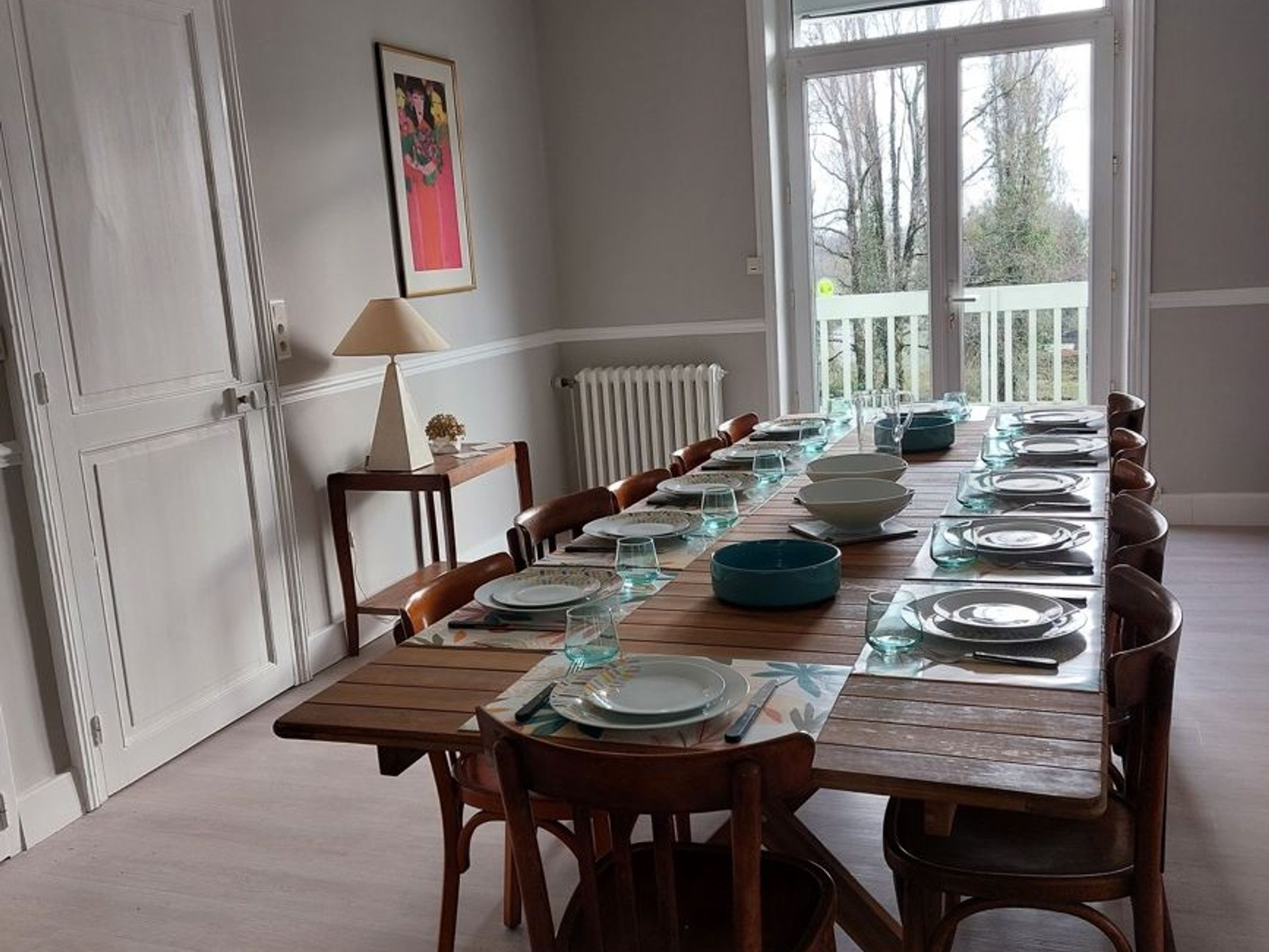 A dining room with a long wooden table set for a meal, flanked by chairs, with a radiator and a window in the background.