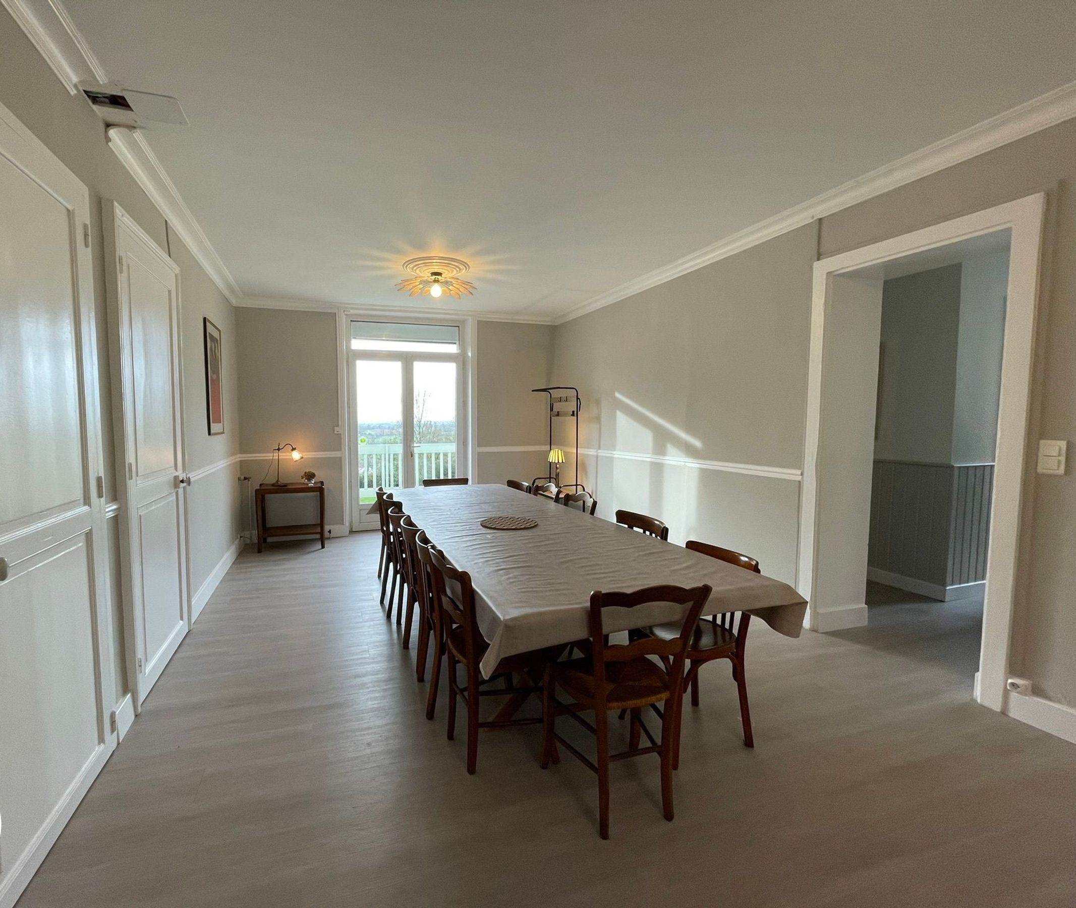 Une salle à manger avec une longue table et des chaises, un ventilateur de plafond et une vue sur l’extérieur, visible à travers une porte vitrée.