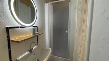 A bathroom with a white sink, a mirror with a light, a shower, and a towel rack.