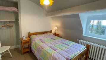 A bedroom with a wooden bed, a bedside table with a lamp, a chair, a wardrobe, and a window with a view.