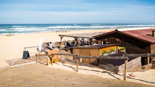 Een houten gebouw aan het strand, mogelijk een restaurant of café, met uitzicht op een zandstrand en de oceaan op de achtergrond.