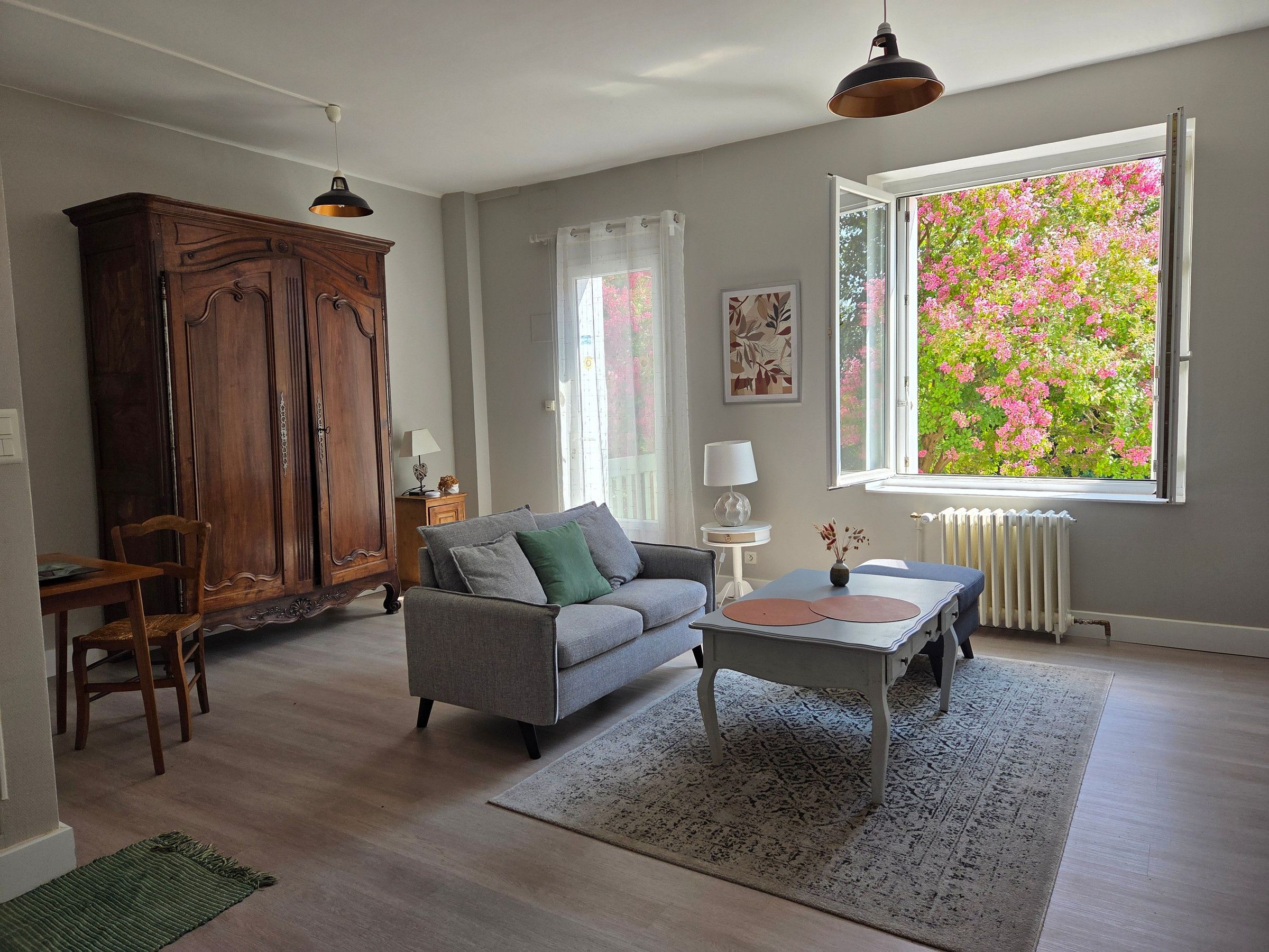A living room with a sofa, a wooden cabinet, a dining table with chairs, a small table with a lamp, and a window with a view of pink flowers.