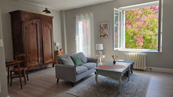 A living room with a sofa, a wooden cabinet, a dining table with chairs, a small table with a lamp, and a window with a view of pink flowers.