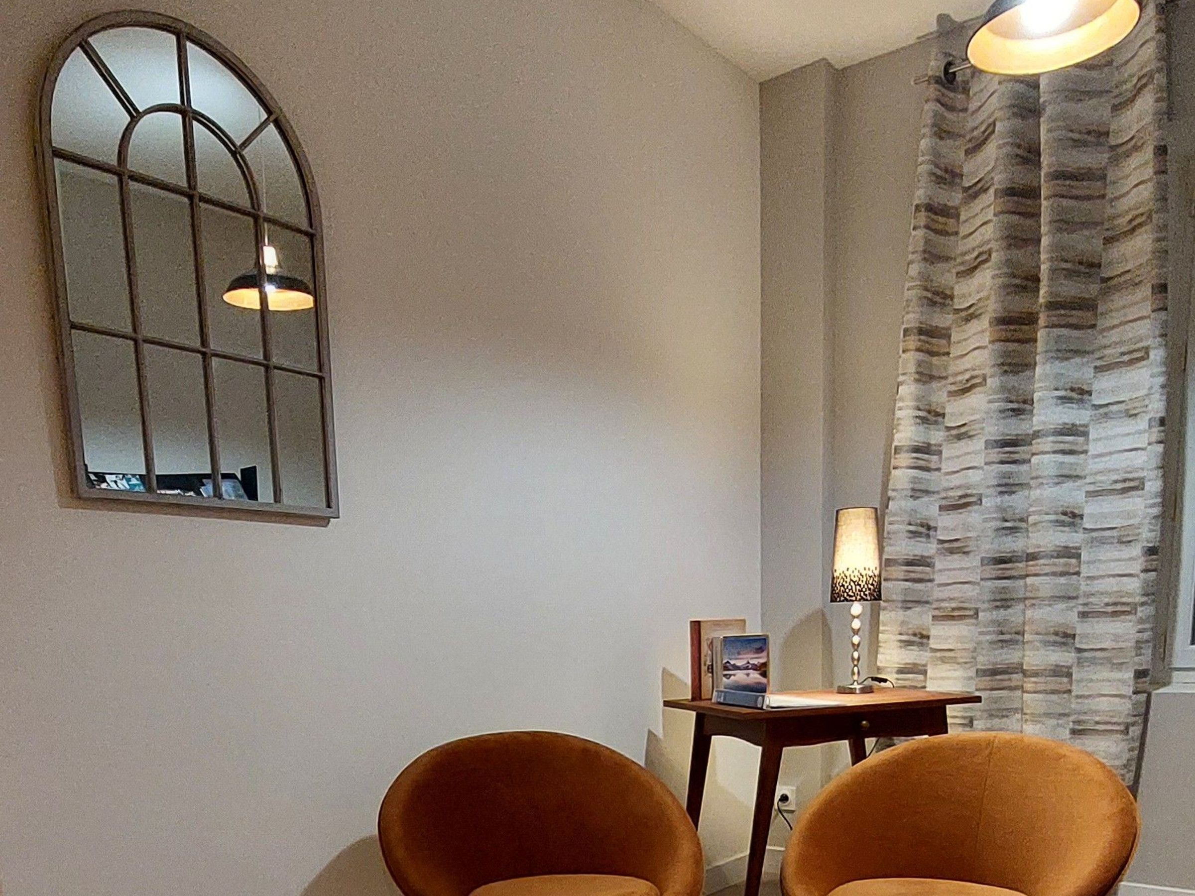 A room with a large arched mirror, two chairs, a small table with a lamp, and a patterned curtain.