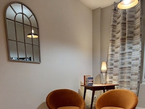 A room with a large arched mirror, two chairs, a small table with a lamp, and a patterned curtain.