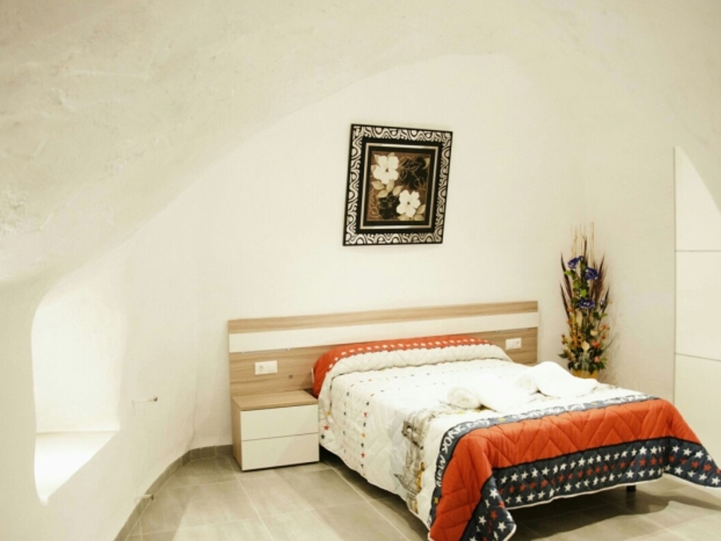 Een slaapkamer met een houten hoofdbord, een wit bed met een geborduurde oranje en blauwe spreideken, een nachtkastje en een aan de muur bevestigd fotolijstje.