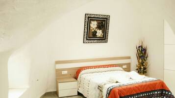 Een slaapkamer met een houten hoofdbord, een wit bed met een geborduurde oranje en blauwe spreideken, een nachtkastje en een aan de muur bevestigd fotolijstje.
