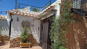 Un pati amb un paviment de rajoles, una banca de fusta, una planta en test i un edifici amb teulada de rajoles i parets de pedra.