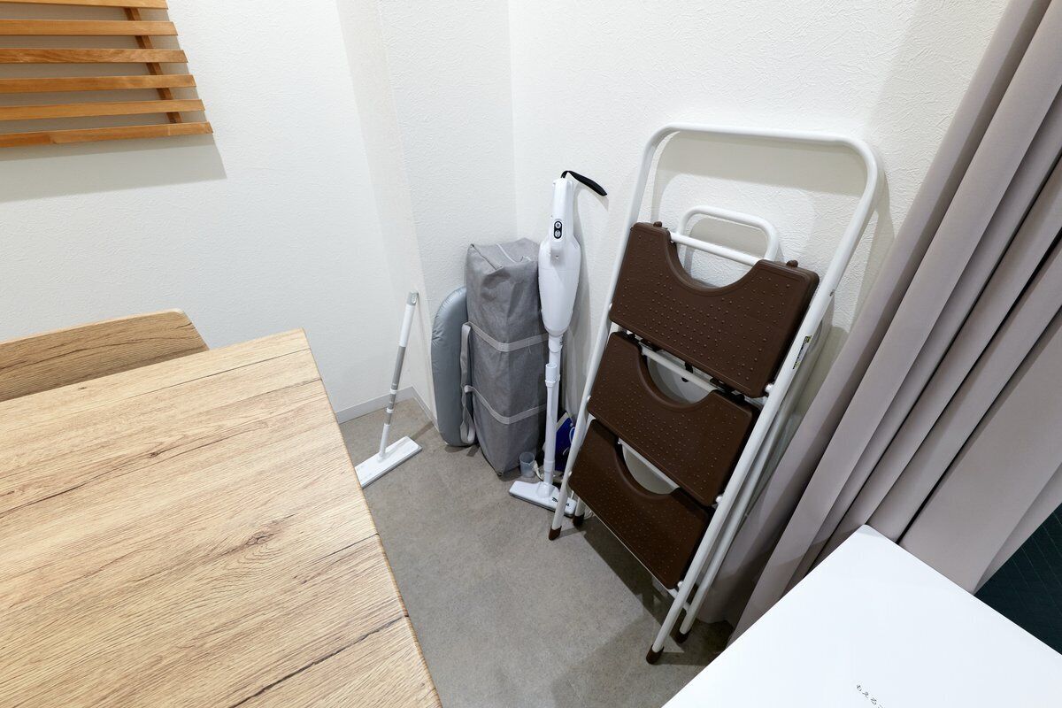 Une petite pièce avec une table en bois, une chaise blanche, une chaise marron, un radiateur blanc et une valise grise.