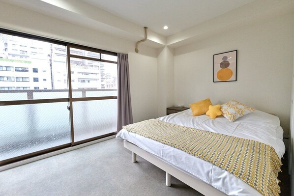 Un dormitorio con un ventanal grande, una cama con ropa de cama blanca y una manta estampada, y una obra de arte enmarcada en la pared.